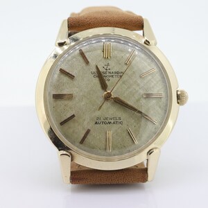 Pu&ograve; includere: Un orologio da polso dorato con quadrante bianco e lancette nere. Il quadrante dell'orologio ha indici orari in numeri romani e il testo "ULYSSE NARDIN CHRONOMETER CO" e "21 JEWELS AUTOMATIC".