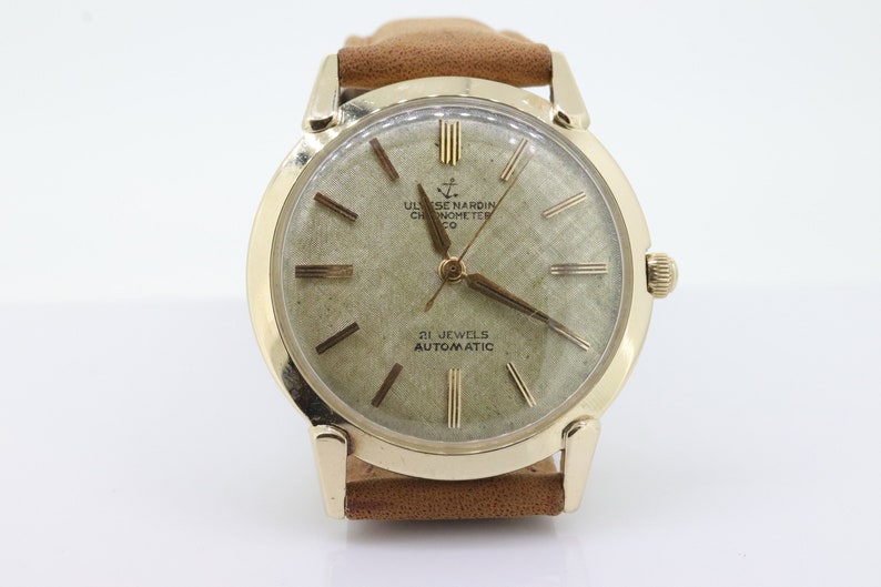 Pu&ograve; includere: Un orologio da polso dorato con quadrante bianco e lancette nere. Il quadrante dell'orologio ha indici orari in numeri romani e il testo "ULYSSE NARDIN CHRONOMETER CO" e "21 JEWELS AUTOMATIC".