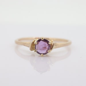 May include: A gold ring featuring a round, light purple gemstone. The ring has a delicate band and decorative gold accents on either side of the gemstone. The ring is set against a white background.