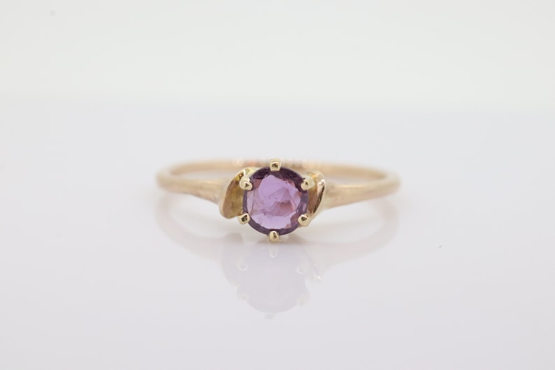 May include: A gold ring with a round, light purple gemstone. The ring has a slender band and decorative gold features either side of the gemstone. The ring is set against a white background.