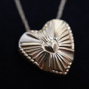 May include: A gold heart-shaped pendant necklace with a smaller heart design in the center. The pendant has a textured, sunburst-like pattern.