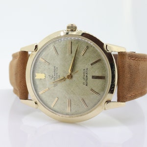 Pu&ograve; includere: Un orologio da polso dorato con quadrante bianco e lancette nere. L'orologio ha un cinturino in pelle marrone e dice "21 Jewels Automatic" e "G. de Marin Chronometer Co."