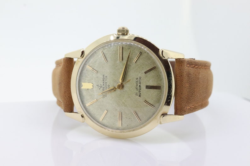 Pu&ograve; includere: Un orologio da polso dorato con quadrante bianco e lancette nere. L'orologio ha un cinturino in pelle marrone e dice "21 Jewels Automatic" e "G. de Marin Chronometer Co."