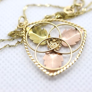 May include: Gold-tone necklace with a heart-shaped pendant. The pendant features three leaf designs in gold, rose, and green tones, encircled by gold rings and a twisted rope border. The chain is a delicate, twisted design.