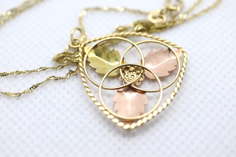 May include: Gold-tone necklace with a heart-shaped pendant. The pendant features three leaf designs in gold, rose, and green tones, encircled by gold rings and a twisted rope border. The chain is a delicate, twisted design.