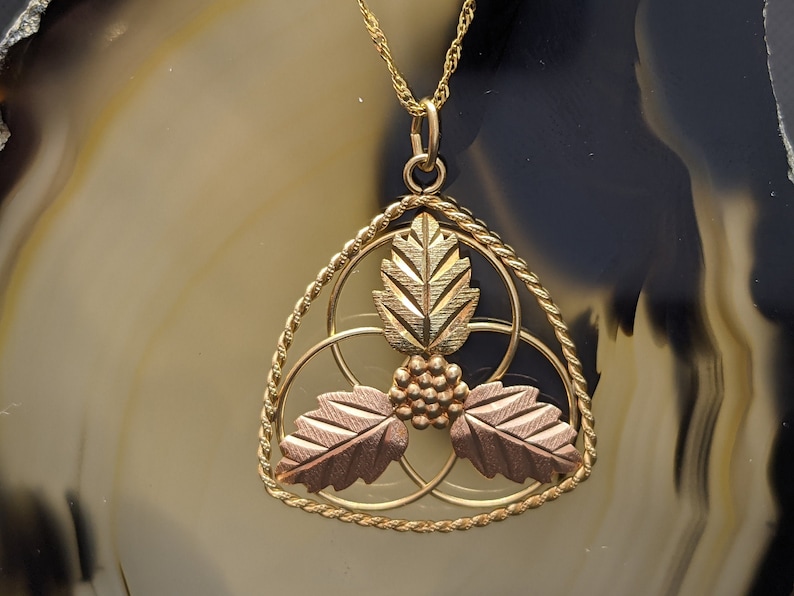 May include: Gold-tone pendant necklace featuring a triangular design with three leaves and a cluster of grapes. The leaves have a two-tone finish, with gold and rose gold accents. The necklace has a delicate gold chain.