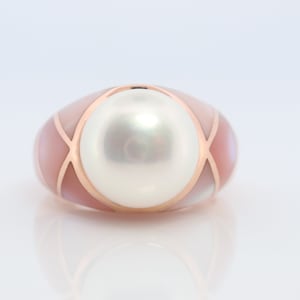 Könnte beinhalten: Ein Ring mit einem großen weißen Perlmutt, das in eine rosafarbene und goldene Fassung gefasst ist. Die Fassung besteht aus Perlmutt und hat ein geometrisches Design.