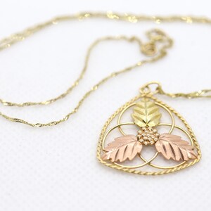 May include: A gold-toned necklace with a triangular pendant featuring three leaves in gold, rose, and yellow hues. The pendant has a delicate, openwork design with a small cluster of beads at the center. The chain is a fine, twisted rope style.