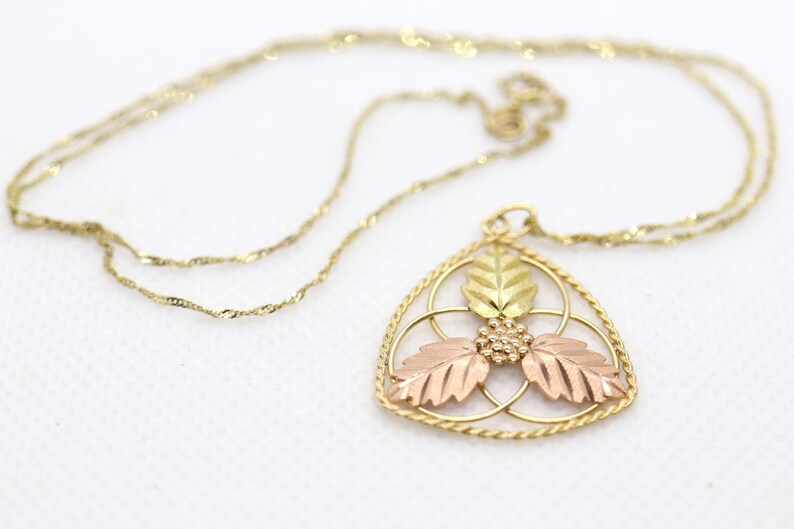 May include: A gold-toned necklace with a triangular pendant featuring three leaves in gold, rose, and yellow hues. The pendant has a delicate, openwork design with a small cluster of beads at the center. The chain is a fine, twisted rope style.