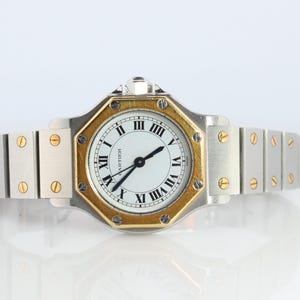 Könnte beinhalten: Eine zweifarbige Armbanduhr mit einem silberfarbenen Edelstahl-Gliederarmband mit goldfarbenen Schraubenakzenten. Das runde weiße Zifferblatt zeigt schwarze römische Ziffern und schwarze Zeiger, wobei das Cartier-Logo auf dem Zifferblatt aufgedruckt ist.