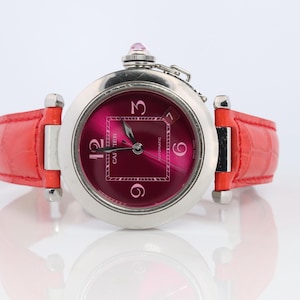 May include: A silver-toned wristwatch with a vibrant red leather band and a deep pink watch face. The watch features a square dial with the numbers 3, 6, 9, and 12. A small pink gem sits atop the watch.