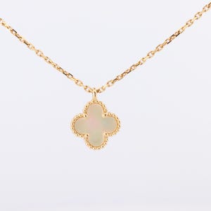 May include: A gold chain necklace featuring a four-leaf clover pendant. The pendant has a smooth, iridescent white inlay surrounded by a beaded gold border. The delicate chain connects to the top of the clover charm, creating a simple, elegant piece.
