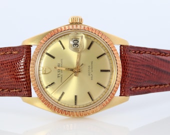 May include: A gold-tone wristwatch with a brown leather band. The watch face is gold with black hands and markers. The watch has a date window at the 3 o'clock position and the text "TUDOR PRINCE OYSTERDATE" and "ROTOR SELF WINDING" on the face.