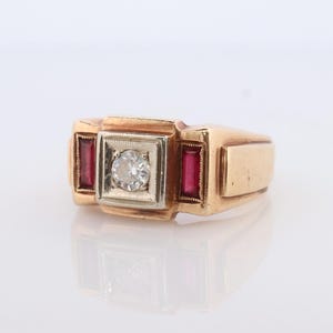 Puede incluir: Un anillo de oro con un diamante central y dos piedras rojas rectangulares. El anillo tiene un diseño rectangular con un engaste cuadrado para el diamante y piedras rojas a cada lado. El anillo está sobre un fondo blanco.