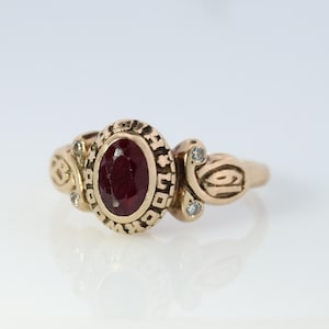 May include: A gold ring with a red oval gemstone and two small diamonds. The ring has a detailed design with a pattern around the gemstone.