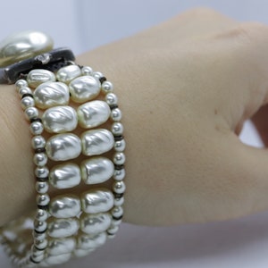 May include: A white pearl bracelet with a black band. The bracelet is made of multiple strands of pearls and has a large pearl at the clasp.