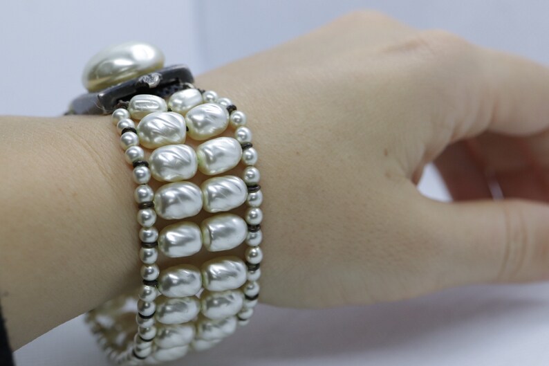 May include: A white pearl bracelet with a black band. The bracelet is made of multiple strands of pearls and has a large pearl at the clasp.