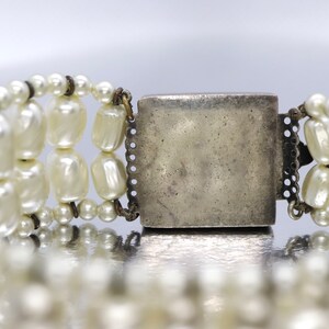 May include: A silver clasp with a decorative design, attached to a multi-strand pearl bracelet.