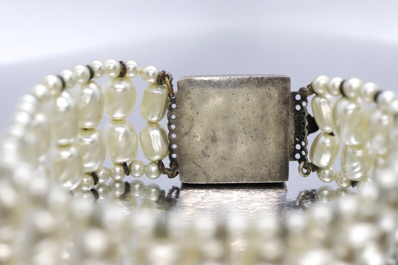 May include: A silver clasp with a decorative design, attached to a multi-strand pearl bracelet.