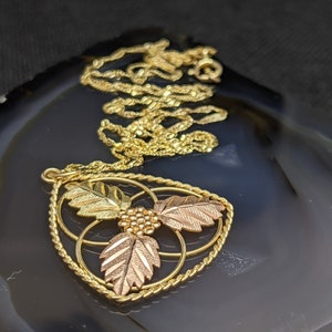 May include: Gold-tone necklace with a pendant featuring three leaves in gold and rose gold tones, and a cluster of small gold beads. The pendant is framed by a twisted gold rope design. The necklace rests on a dark, patterned surface.