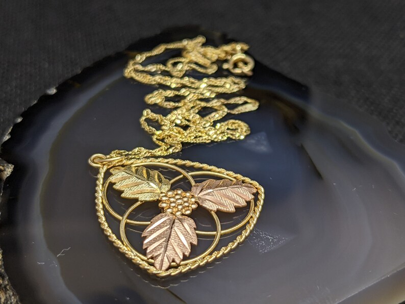 May include: Gold-tone necklace with a pendant featuring three leaves in gold and rose gold tones, and a cluster of small gold beads. The pendant is framed by a twisted gold rope design. The necklace rests on a dark, patterned surface.