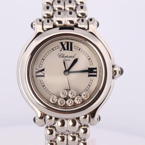 May include: A silver-toned Chopard Geneve wristwatch with a round face, Roman numeral markers, and black hands. The dial features seven loose, circular-cut diamonds that move freely within the case. It has a polished metal link bracelet.