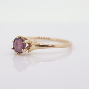 May include: A gold ring with a round, purple gemstone. The gemstone is secured by six prongs. The ring's band is slender and polished. The ring is set against a white backdrop.