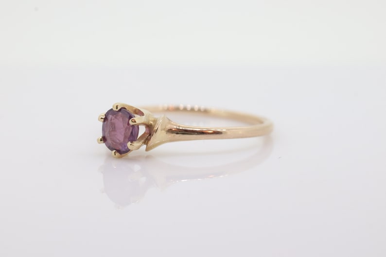 May include: A gold ring with a round, purple gemstone. The gemstone is secured by six prongs. The ring's band is slender and polished. The ring is set against a white backdrop.