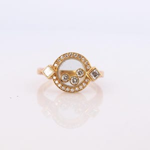 May include: A gold ring featuring a circular center with a clear glass pane containing three small, loose, round-cut diamonds. The circular frame is bordered by tiny pavé-set diamonds, with a square-cut diamond on each side of the central setting.