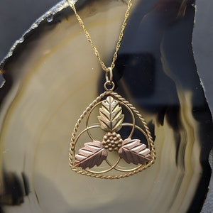 May include: A gold-toned pendant necklace featuring a trinity design with three leaves and a cluster of berries. The leaves are in shades of gold, rose, and green. The necklace is displayed on a dark, patterned background.