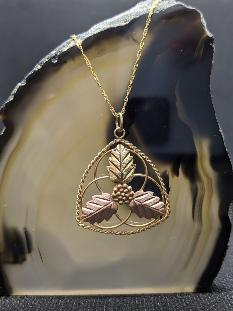 May include: A gold-toned pendant necklace featuring a trinity design with three leaves and a cluster of berries. The leaves are in shades of gold, rose, and green. The necklace is displayed on a dark, patterned background.