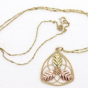 May include: Gold-tone necklace with a pendant featuring a leaf design in gold, rose, and yellow tones. The pendant is a triangular shape with a detailed floral pattern. The chain is a delicate, twisted rope style.