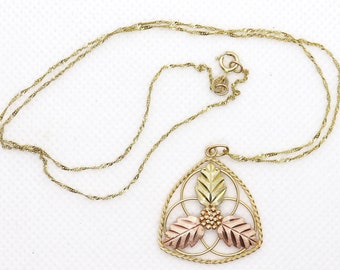 May include: Gold-tone necklace with a pendant featuring a leaf design in gold, rose, and yellow tones. The pendant is a triangular shape with a detailed floral pattern. The chain is a delicate, twisted rope style.