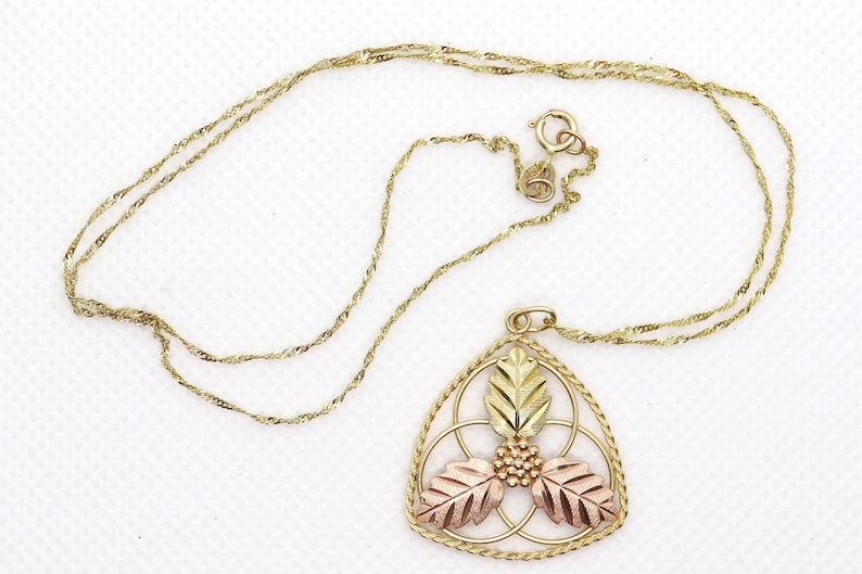 May include: Gold-tone necklace with a pendant featuring a leaf design in gold, rose, and yellow tones. The pendant is a triangular shape with a detailed floral pattern. The chain is a delicate, twisted rope style.