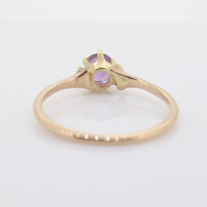 May include: A gold ring featuring a round, light purple gemstone. The ring has a slender band and decorative detailing around the gemstone setting. The ring is set against a white background.