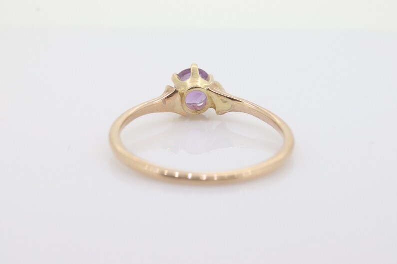 May include: A gold ring featuring a round, light purple gemstone. The ring has a slender band and decorative detailing around the gemstone setting. The ring is set against a white background.