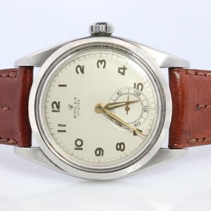 May include: A vintage Rolex Oyster watch with a silver-colored case and a cream-colored dial. The watch features gold-colored hands and numerals, and a small seconds dial. It has a brown leather strap.