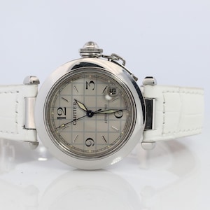May include: A silver-toned Cartier Pasha C automatic watch with a white leather strap. The watch face is silver with black numerals and hands. The watch has a date display at the 3 o'clock position. The white strap is textured.