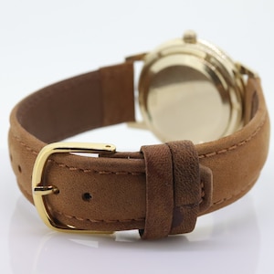 Pu&ograve; includere: Un orologio da polso in oro con un cinturino in pelle marrone. L'orologio ha un quadrante rotondo e una fibbia dorata.