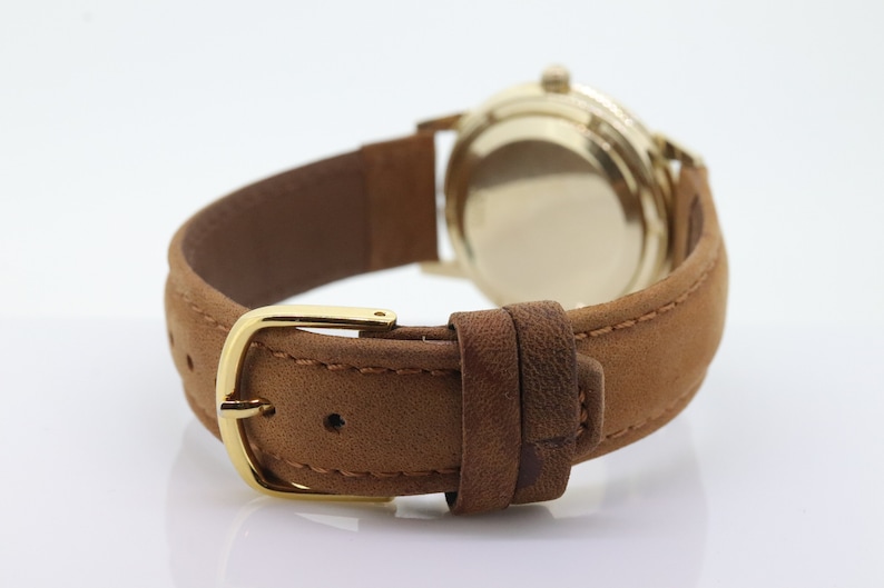 Pu&ograve; includere: Un orologio da polso in oro con un cinturino in pelle marrone. L'orologio ha un quadrante rotondo e una fibbia dorata.