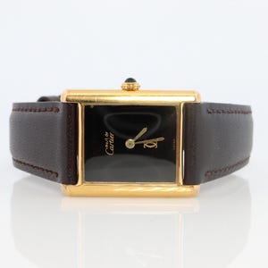 May include: A gold-toned rectangular watch with a black face and brown leather band. The watch face has gold hands and the words "Must de Cartier" in gold lettering. The watch band is dark brown with visible stitching.