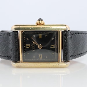 May include: A gold-toned rectangular wristwatch with a black dial and Roman numeral hour markers. The watch features a black leather band and a small crown with a black stone. The watch face has the brand name "Cartier" printed on it.