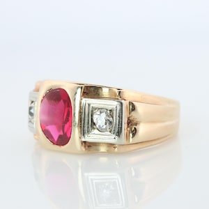 Puede incluir: Un anillo dorado con una gran gema de rubí ovalada y dos diamantes más pequeños. El anillo tiene un diseño clásico con un engaste rectangular para los diamantes y un acabado pulido. El anillo se muestra sobre una superficie blanca.