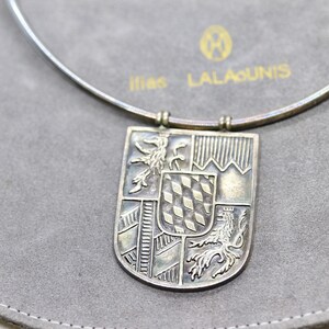 May include: A silver necklace with a pendant featuring a detailed coat of arms design. The pendant is shaped like a shield and has four sections, each with a different design. The necklace is on a gray background with the text "1186 LALAOUNIS".