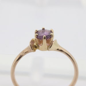 May include: A gold ring featuring a round, light purple gemstone. The ring has a slender band and an ornate setting that holds the gemstone. The ring is set against a white background.