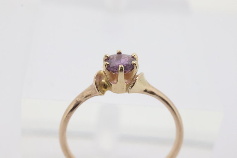 May include: A gold ring featuring a round, light purple gemstone. The ring has a slender band and an ornate setting that holds the gemstone. The ring is set against a white background.