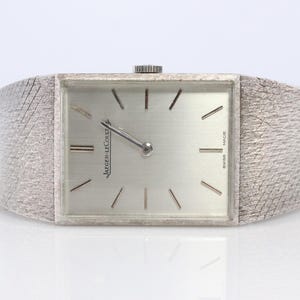 May include: A silver-toned rectangular wristwatch with a textured band. The watch face is silver with black markers and hands. The brand name "JAEGER-LECOULTRE" is printed on the watch face. The watch is marked "SWISS MADE".