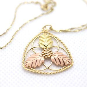 May include: Gold-toned necklace with a pendant featuring a leaf design. The pendant has three leaves in gold, rose gold, and green gold, with a cluster of small gold beads. The chain is a delicate, twisted design.