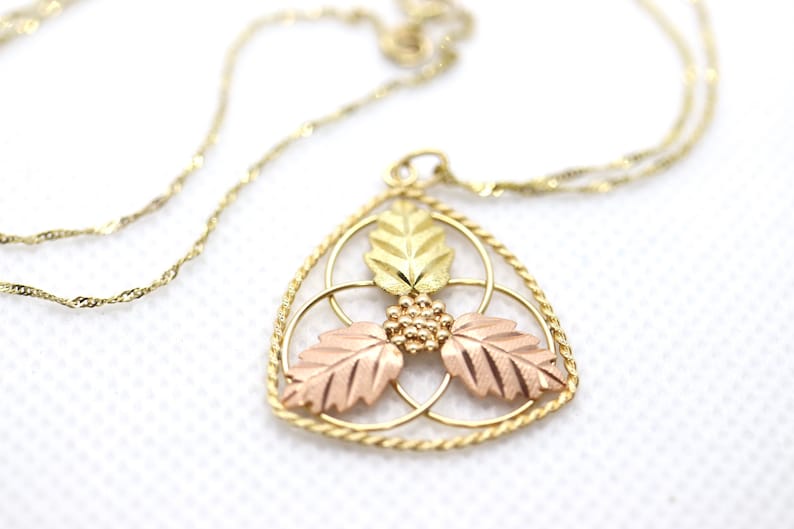 May include: Gold-toned necklace with a pendant featuring a leaf design. The pendant has three leaves in gold, rose gold, and green gold, with a cluster of small gold beads. The chain is a delicate, twisted design.