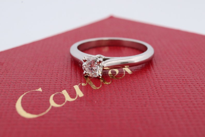 May include: A solitaire diamond engagement ring with a round brilliant cut diamond set in a platinum band. The ring is displayed on a red box with the word 'Cartier' printed in gold lettering.